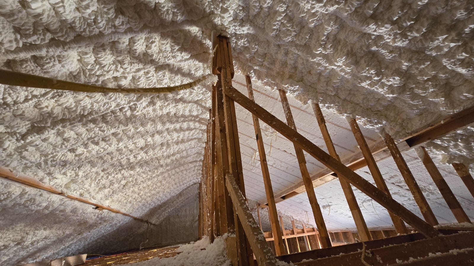 Spray foam insulation applied to attic roofline framing during installation, showing rafters and raw sprayed texture before trimming